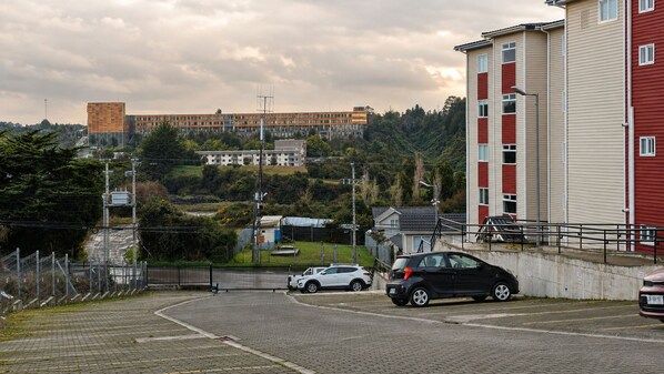 A parking area with cars, a building with red and white siding, and a distant view of a larger complex with multiple buildings.