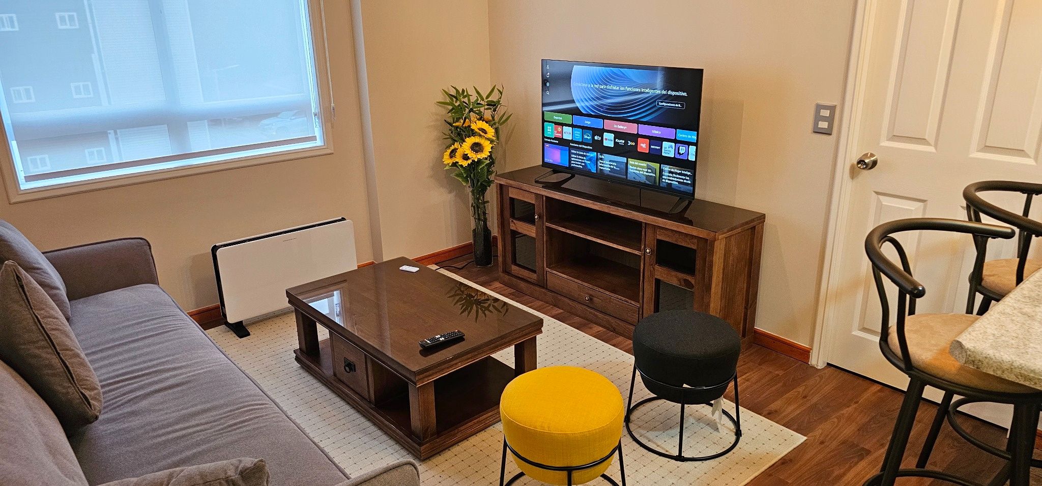 A living room with a sofa, a coffee table, a TV on a wooden stand, a yellow ottoman, and a chair.