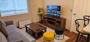 A living room with a sofa, a coffee table, a TV on a wooden stand, a yellow ottoman, and a chair.
