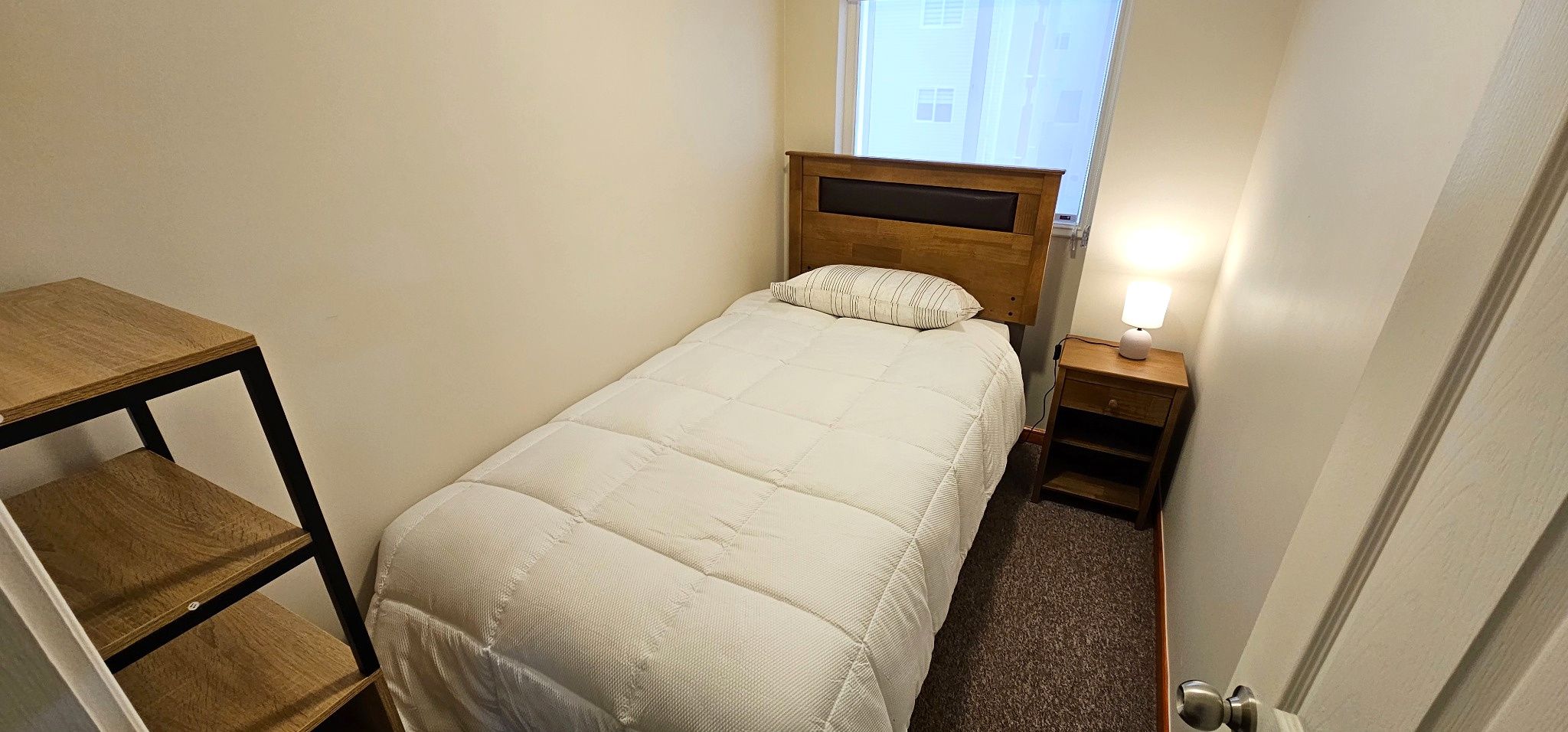 Une petite chambre à coucher, bien rangée, avec un lit, une table de chevet, un bureau et une fenêtre.