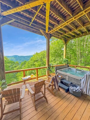 Un terrazzo in legno con vasca idromassaggio, posti a sedere e una vista panoramica su una catena montuosa boscosa.