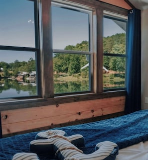Ein Schlafzimmer mit großem Fenster, einem Bett mit blauer Decke und Blick auf einen See und Bäume.