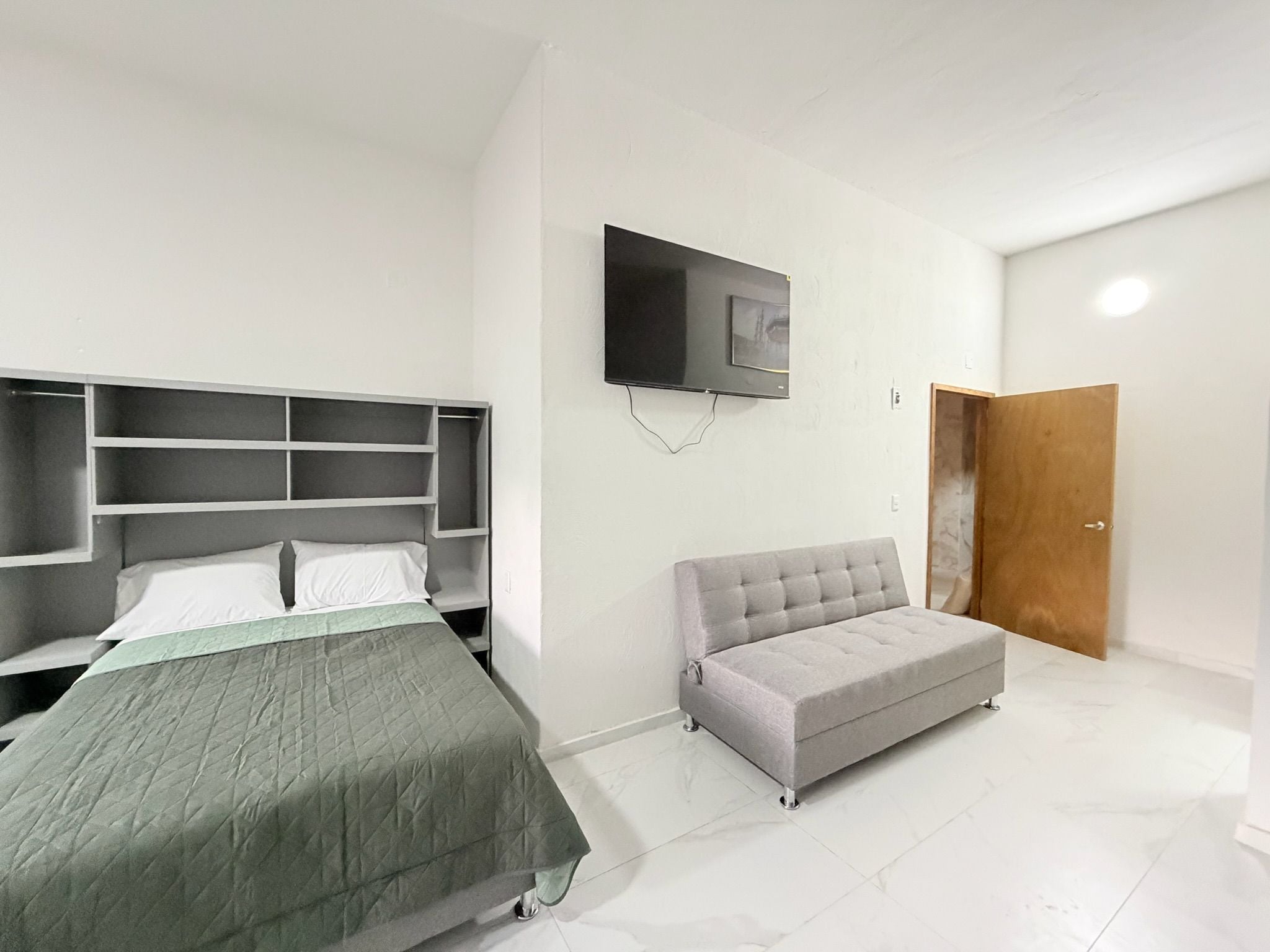 A bedroom with a bed, a grey sofa, a TV mounted on the wall, and a wooden door.