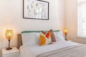 Ein ordentlich bezogenes Bett mit einer gemusterten Decke, zwei Nachttischlampen und einem gerahmten Pflanzenbild an der Wand.