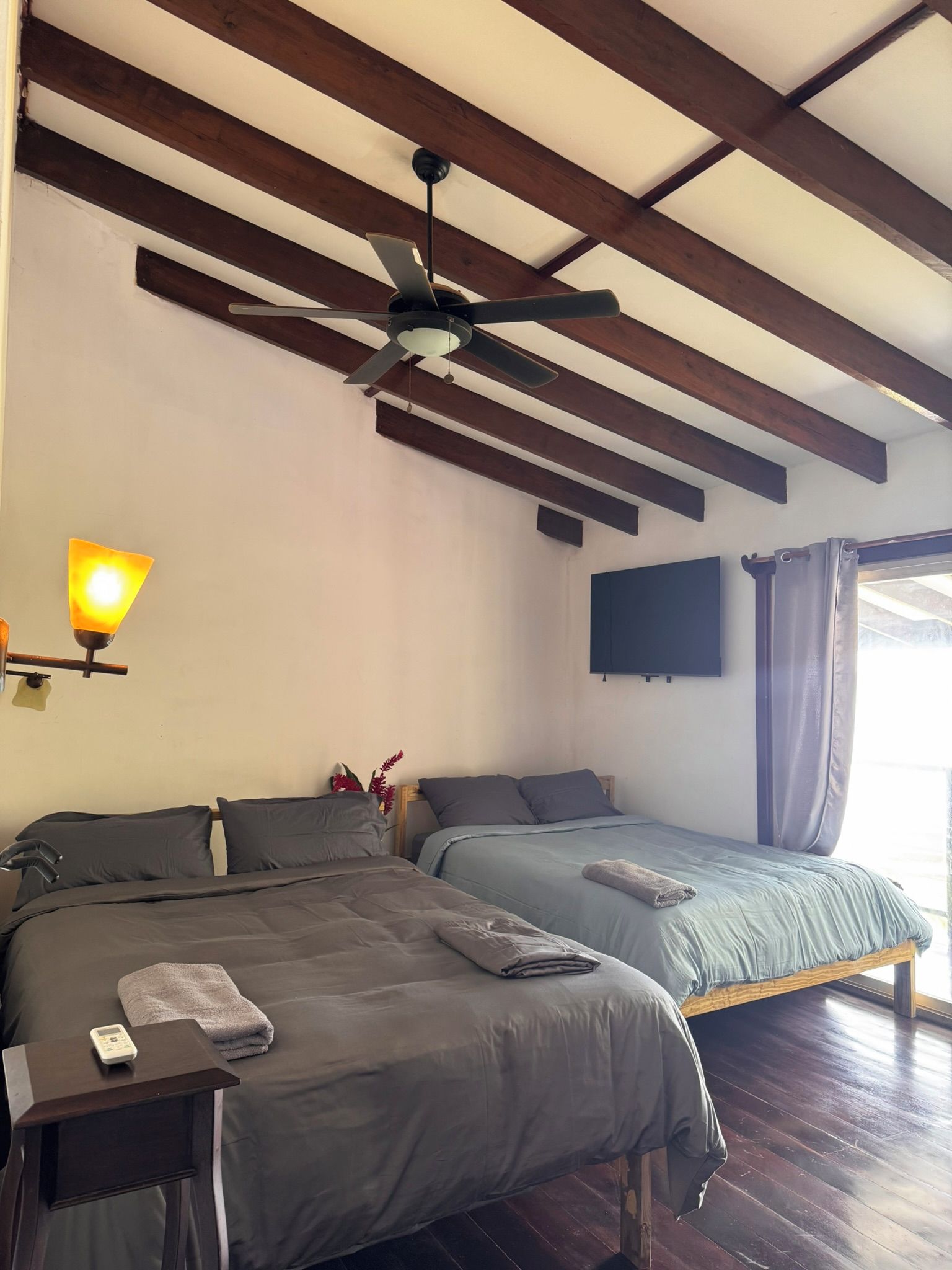 Un dormitorio con dos camas, ventilador de techo, televisor montado y mesita de noche con lámpara.