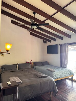 A bedroom with two beds, a ceiling fan, a mounted TV, and a bedside table with a lamp.