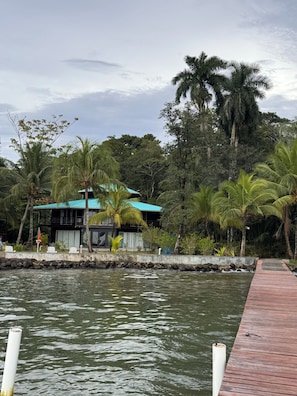 Una propiedad frente al agua con un muelle de madera, un edificio con techo azul y exuberante vegetación tropical.