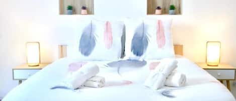 A neatly made bed with pillows and towels, two bedside tables with lamps, and two small wooden shelves with plants.