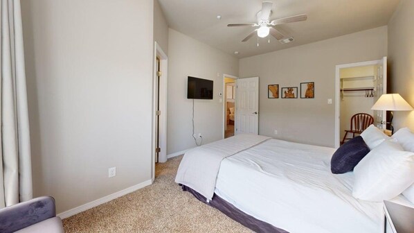 A hotel room with a bed, a ceiling fan, a TV mounted on the wall, and a door leading to another room.