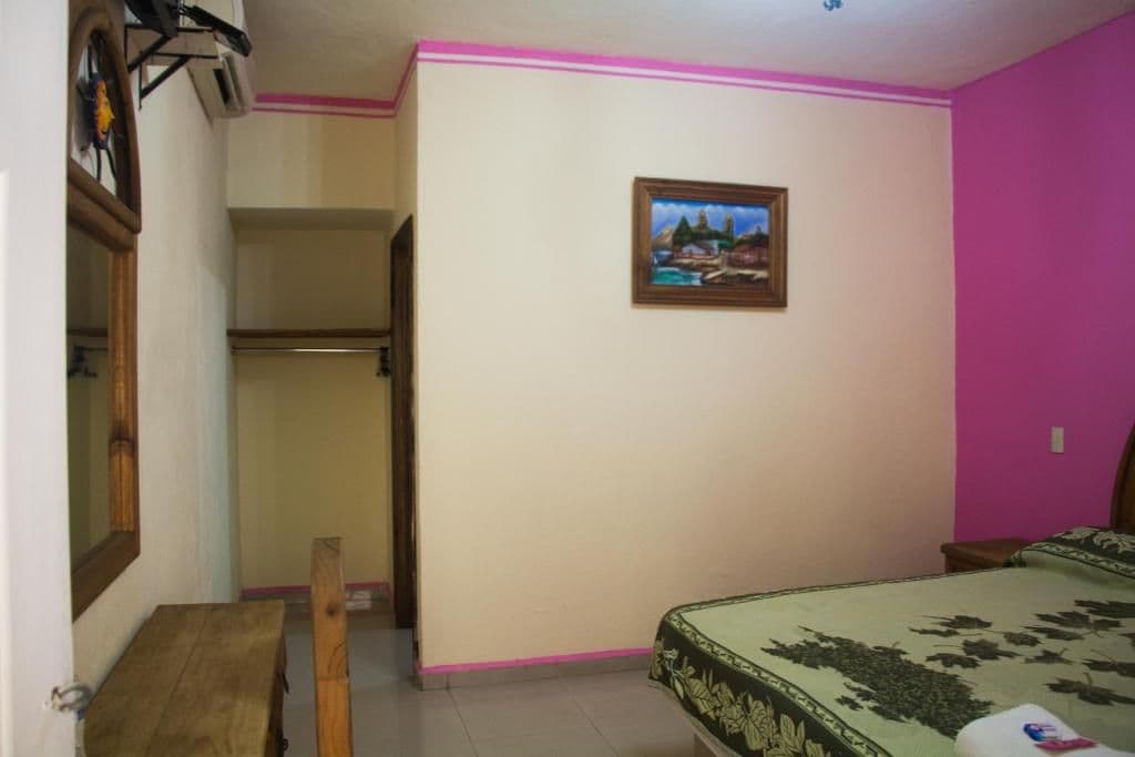 A bedroom with a bed, a chair, a small table, a painting on the wall, and a pink accent wall.