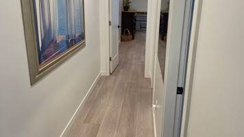 A hallway with a wooden floor, a framed painting on the wall, and a door leading to a room.