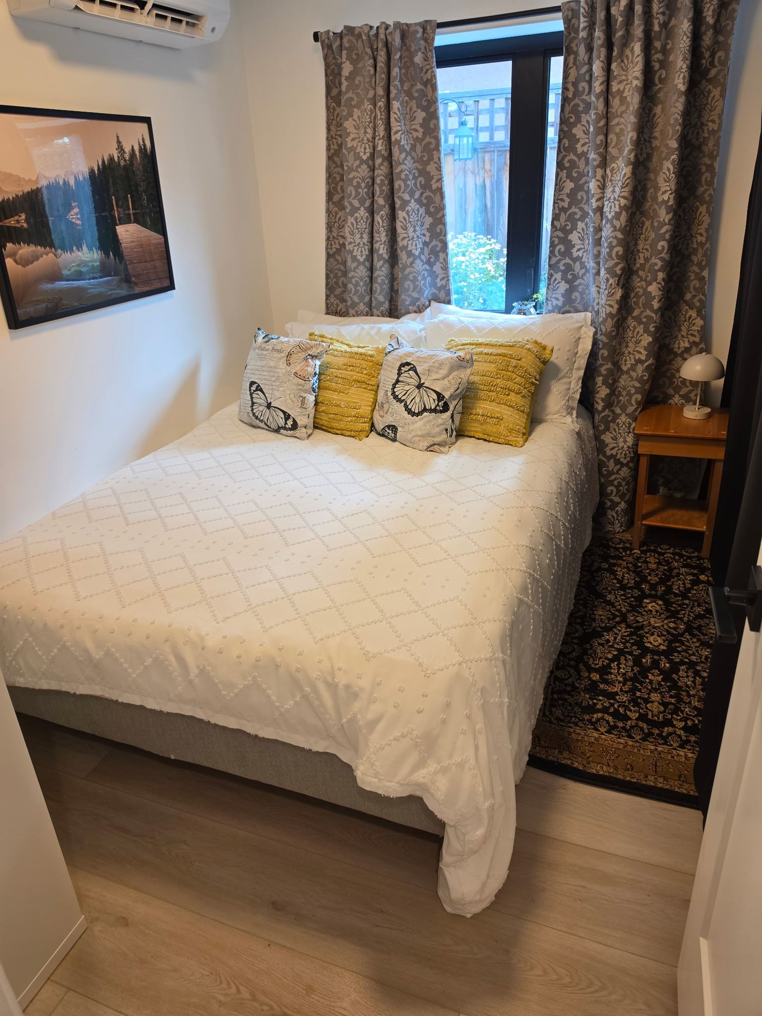 Een slaapkamer met een bed, een raam met gordijnen, een wandairconditioner, een ingelijste foto en een klein nachtkastje met een lamp.