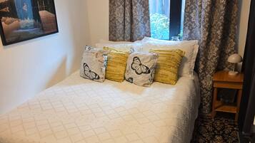 Een slaapkamer met een bed, een raam met gordijnen, een wandairconditioner, een ingelijste foto en een klein nachtkastje met een lamp.