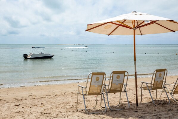 A beach with three lounge chairs under a large umbrella, a boat in the water, and a clear sky.