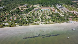 An aerial view of a resort with multiple bungalow-style accommodations, a central building possibly for dining or events, and a beach area with clear water.