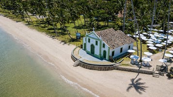 Un bâtiment situé en bord de mer, avec une façade blanche et des éléments décoratifs verts, à côté d’une plage de sable et de palmiers.