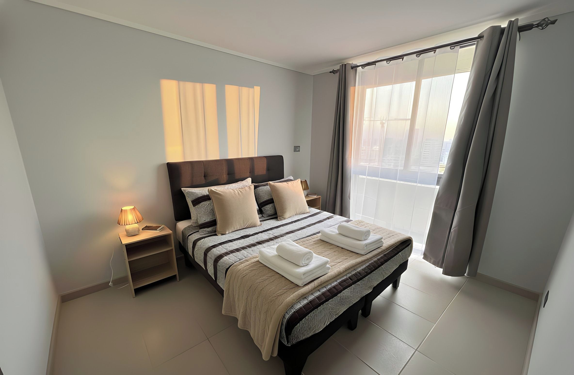 A modern bedroom with a large bed, a bedside table with a lamp, and a view of the city through the window.