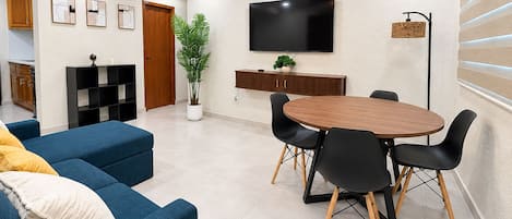 A modern living room with a blue sofa, a wooden dining table with black chairs, a flat-screen TV mounted on the wall, and a ceiling fan.