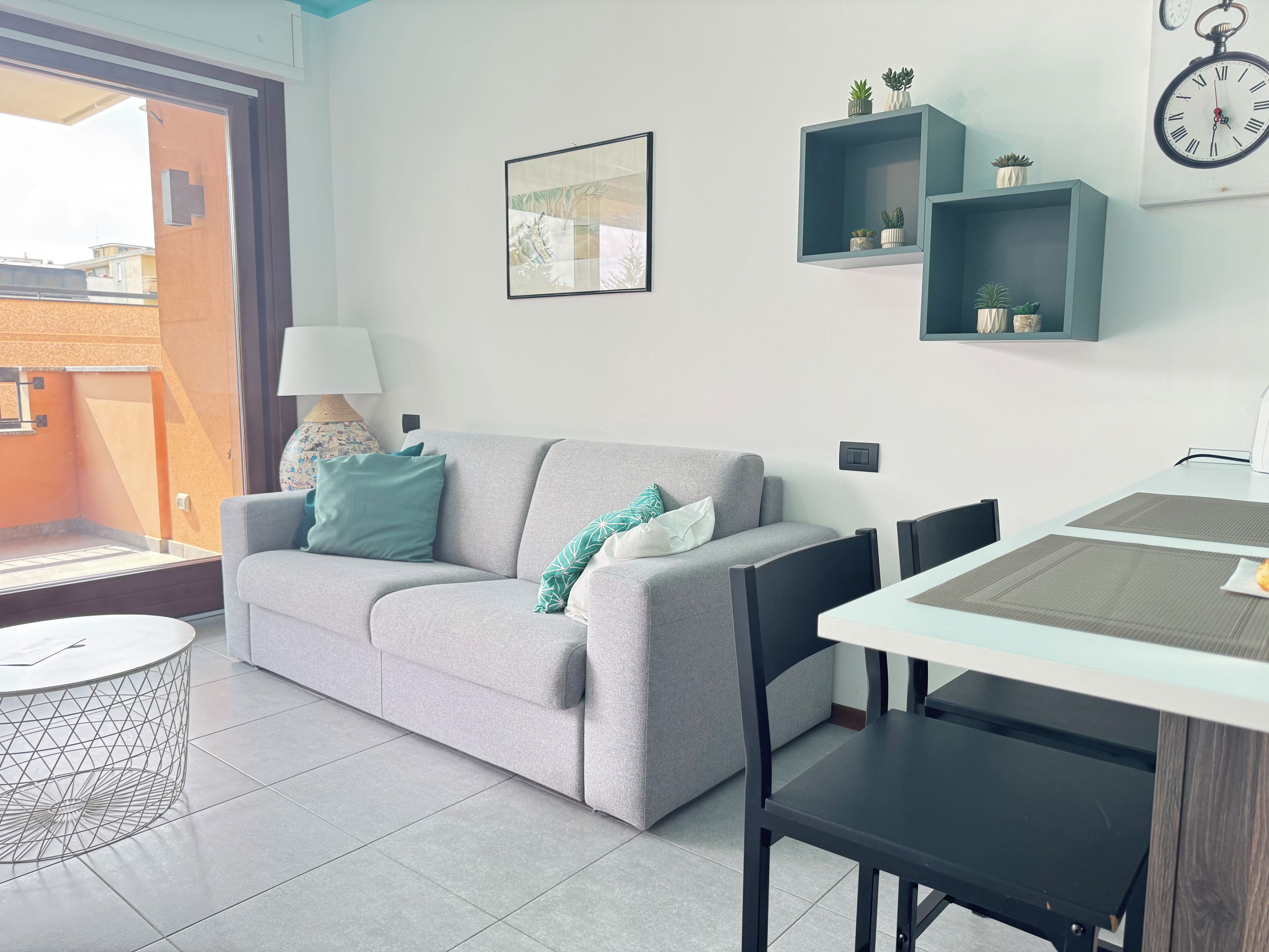Ein modernes Wohnzimmer mit einem grauen Sofa, einem runden Couchtisch und einem Esstisch mit Stühlen. An den Wänden sind Regale mit Pflanzen und eine Wanduhr angebracht.