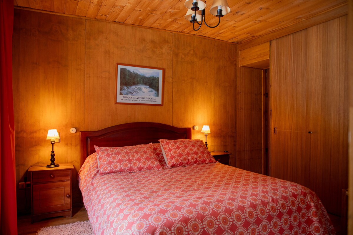 A wooden room with a bed, two bedside lamps, a framed picture on the wall, and a wooden door.