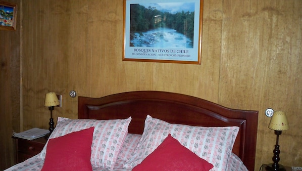 A wooden-paneled room with a bed featuring patterned pillows, a framed picture on the wall, and two bedside lamps. - Casa Verde Hostal Ecologico (Curacautin)