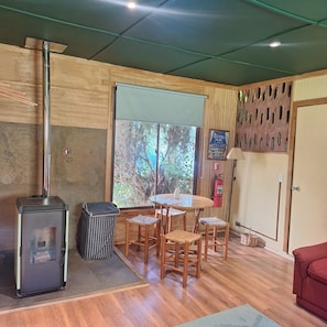 A room with a wood-burning stove, a table with chairs, a fire extinguisher, and a window with a view of trees. - Casa Verde Hostal Ecologico (Curacautin)