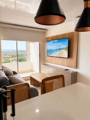 Ein modernes Wohnzimmer mit einem großen, an der Wand befestigten Fernseher, einer Couch, einem Esstisch und Blick auf die Stadt durch große Fenster.
