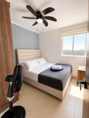 Ein Schlafzimmer mit Bett, Deckenventilator, Fenster und Standventilator.