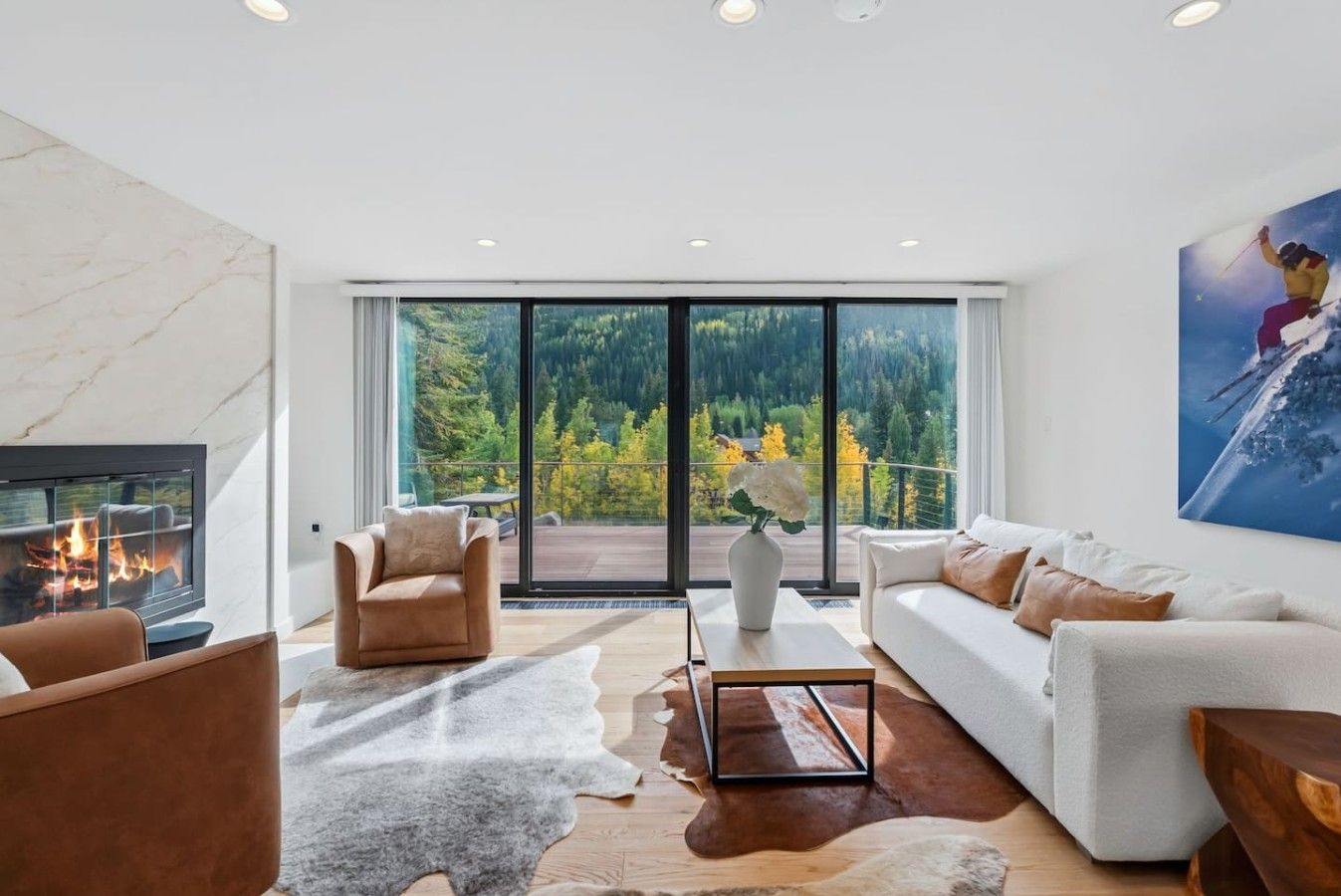 A modern living room with a fireplace, leather armchairs, a white sofa, and a large window overlooking a forest.