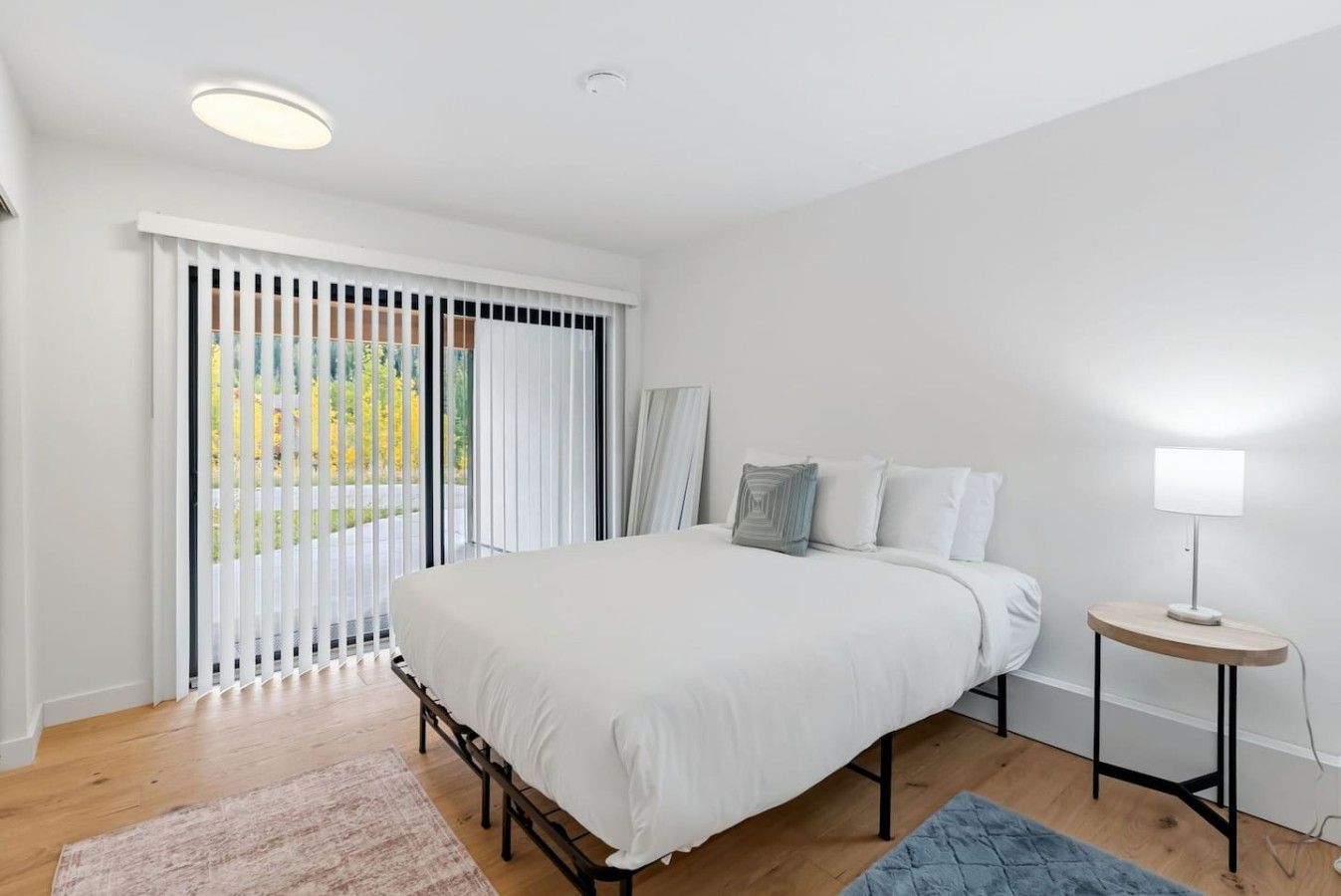 A bedroom with a bed, a bedside table with a lamp, a window with blinds, and a view of the outdoors.