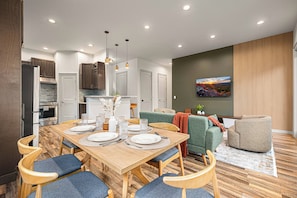 Een moderne keuken met inbouwapparatuur, een eethoek met een houten tafel en blauwe fauteuils, en een zithoek met een groene bank en een grote flatscreen-televisie aan de muur.