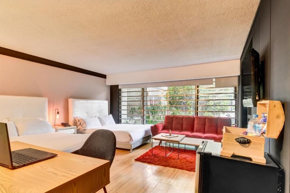 Habitación de hotel moderna con cama, escritorio con ordenador portátil, sofá rojo y ventana grande con vistas a zonas verdes.