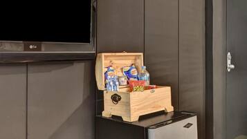 Eine kleine Holzkiste mit Snacks und Getränken steht auf einem schwarzen Gerät, wahrscheinlich einem Minikühlschrank, in einem Raum mit dunkelholzbepflanzten Wänden und einem flachbildfernseher, der darüber angebracht ist.