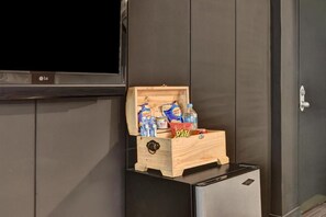 Un pequeño cofre de madera con botanas y bebidas sobre un electrodoméstico negro, probablemente un mini refrigerador, en una habitación con paredes de madera oscura y un televisor de pantalla plana montado en la pared.