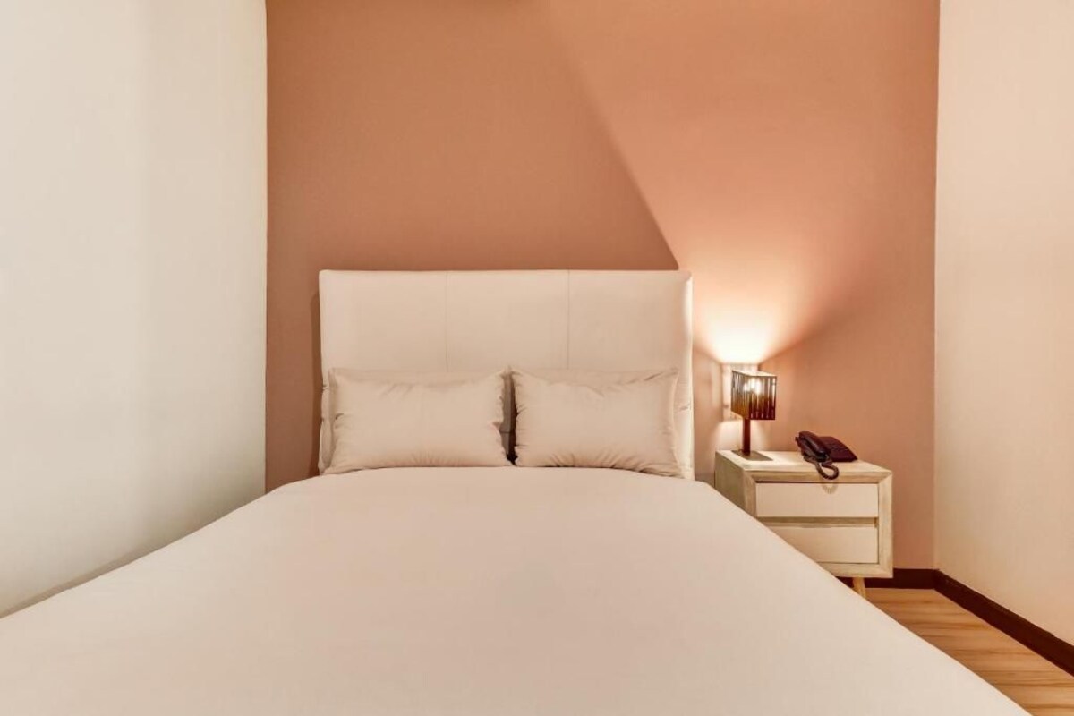 Una cama individual con cabecera, una mesita de noche con lámpara y un teléfono a la derecha.