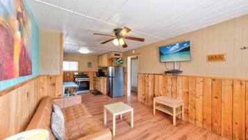 A cozy room with a wooden floor, a sofa, a small table, a ceiling fan, and a television mounted on the wall.