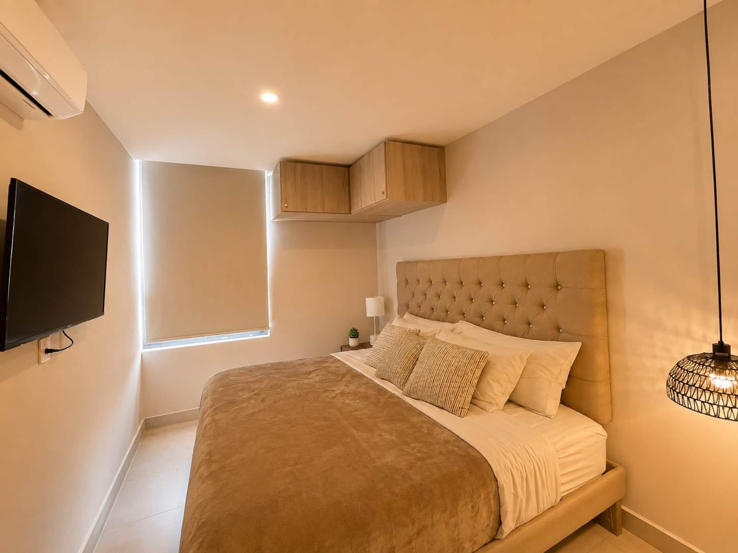 A bedroom with a large bed, a television mounted on the wall, a window with blinds, and a ceiling light fixture.