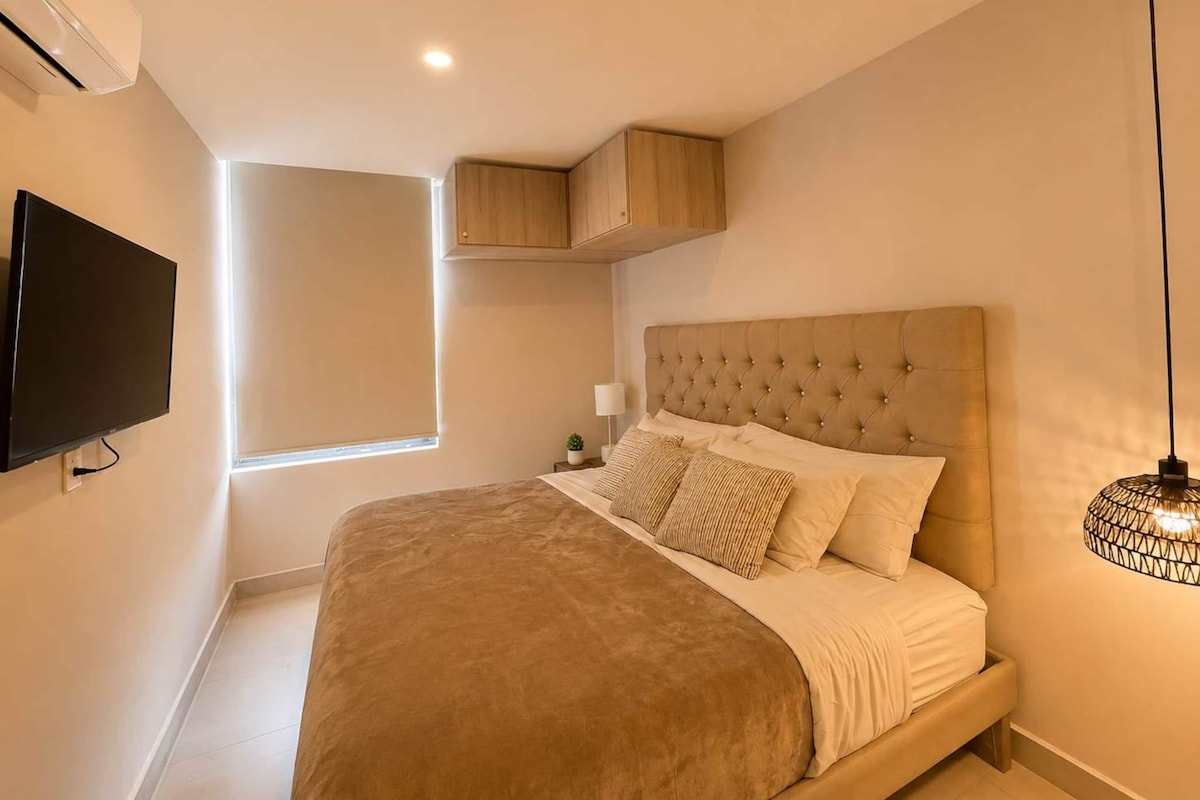 A bedroom with a large bed, a television mounted on the wall, a window with blinds, and a ceiling light fixture.