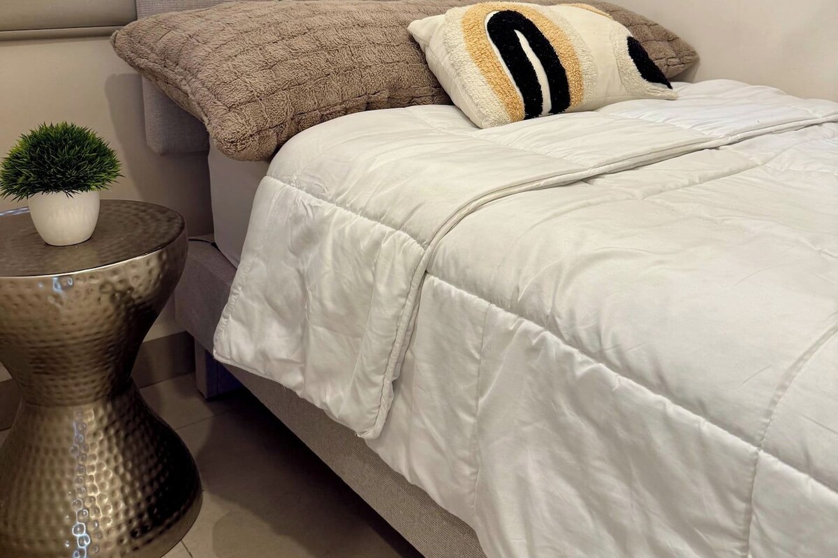 Una cama bien hecha con edredón blanco y dos almohadas, una planta pequeña en maceta sobre una mesita redonda, y un cabecero tapizado en gris.