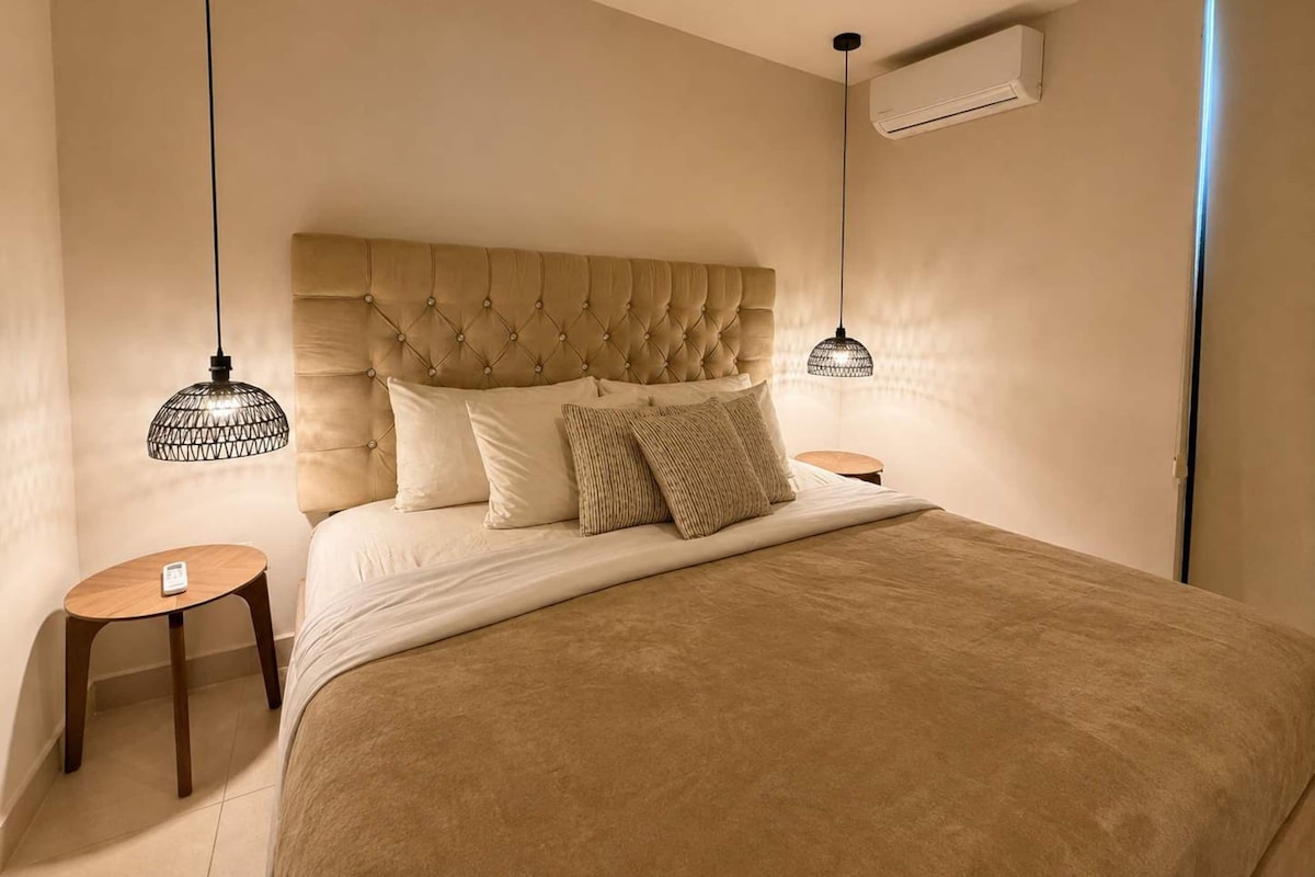 Un dormitorio con cabecera capitonada, dos mesitas de noche, una cama con edredón color beige y lámparas colgantes.