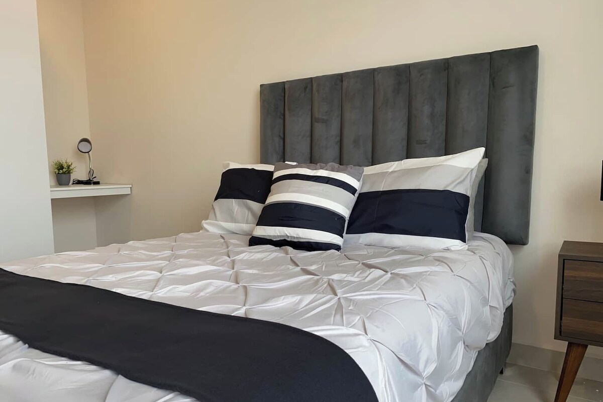 A neatly made bed with a grey upholstered headboard, a bedside table, and a small shelf with a plant.