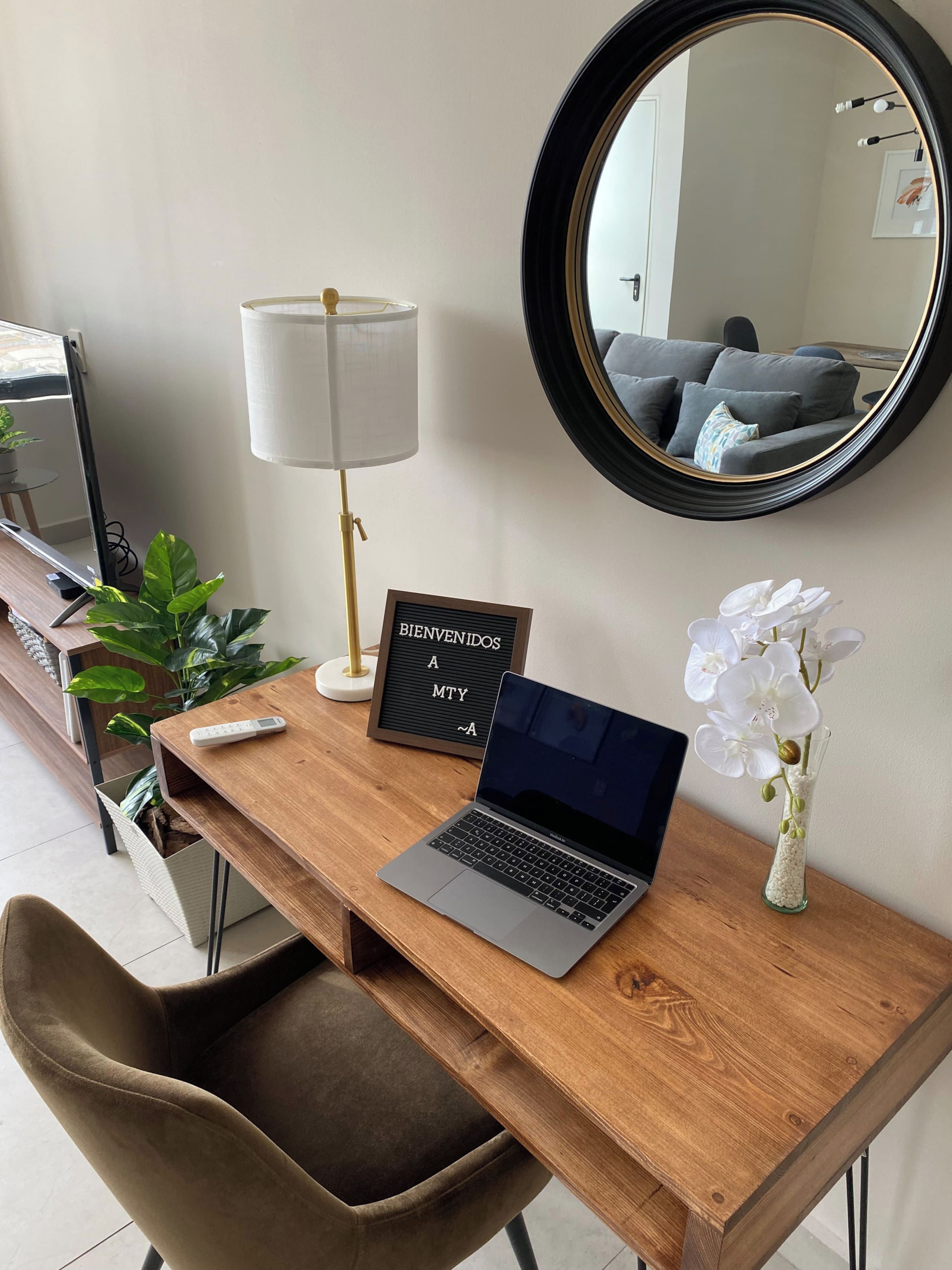 A wooden desk with a laptop, a 'BIENVENIDOS A MTY' sign, a lamp, a vase with white flowers, and a mirror.