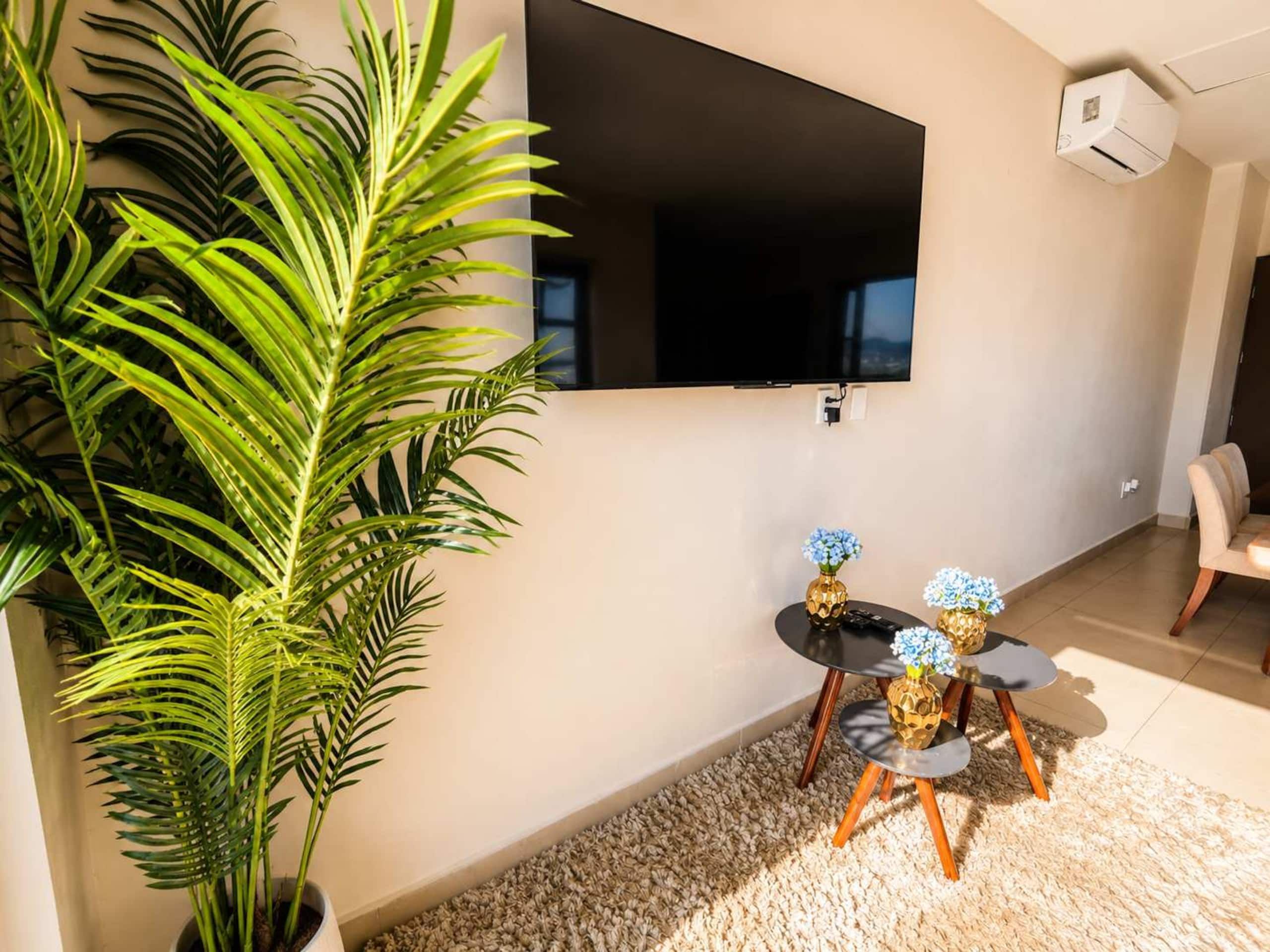 A living room with a flat-screen TV mounted on the wall, a potted plant, a small round table with two vases, and a beige carpet.