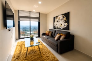 A modern living room with a sofa, a coffee table, and a large window offering a city view. - Arena Vive El Parque Apartamentos  (Monterrey)