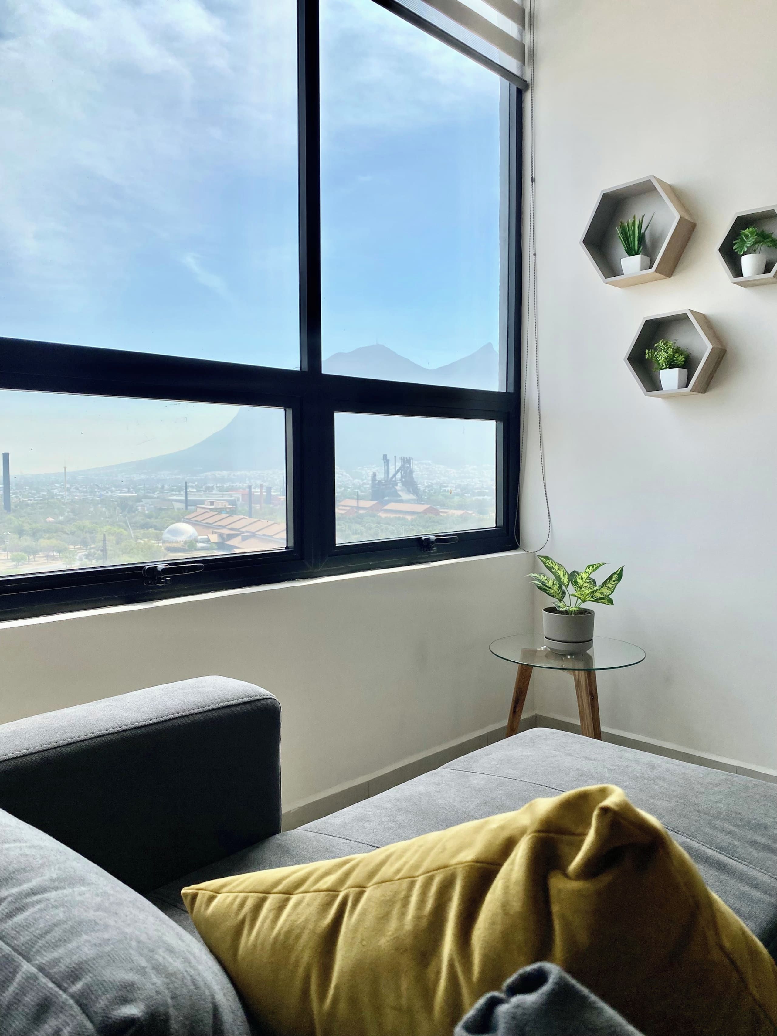 A room with a large window, a grey sofa, a small round table with a plant, and three hexagonal shelves with plants.