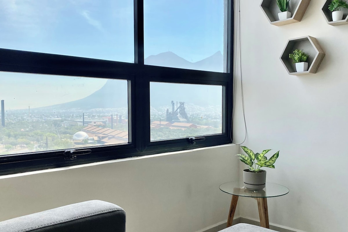A room with a large window, a grey sofa, a small round table with a plant, and three hexagonal shelves with plants.