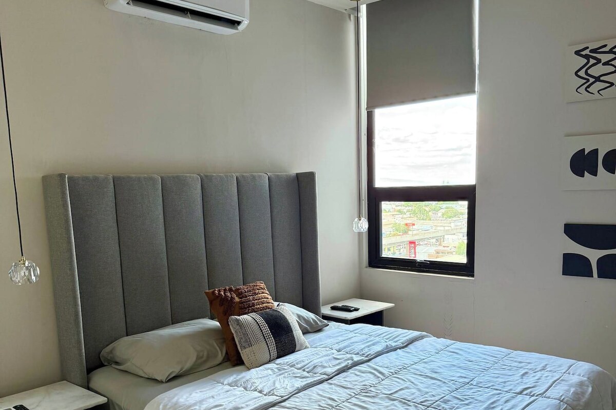 Un dormitorio con cama, mesitas de noche, aire acondicionado, ventana con vistas y una lámpara de pared.