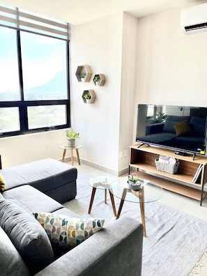 A modern living room with a sofa, a glass coffee table, a TV on a wooden stand, and a view of a mountain through large windows.