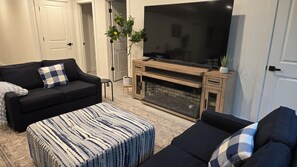 Sala de estar com sofá preto, um banquinho estampado, lareira e uma grande televisão de tela plana.