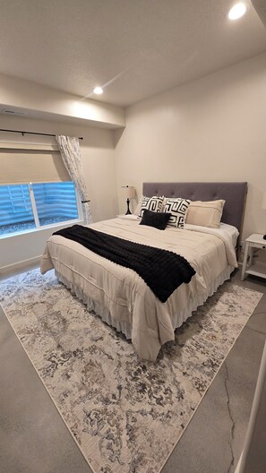 Quarto com cama grande, mesa de cabeceira, janela com cortinas e luminária no teto.